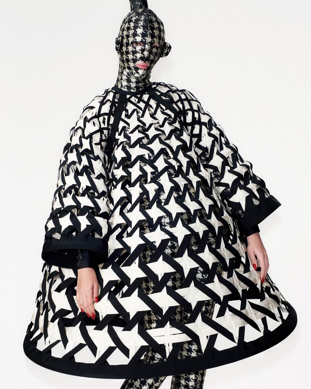 Person in black-and-white star-patterned coat and matching full face covering stands against white backdrop in fashion photograph.