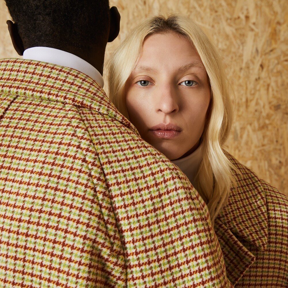 Close-up of a light-skinned woman with blonde hair and a patterned jacket, partially obscured by a Black person in a matching jacket.