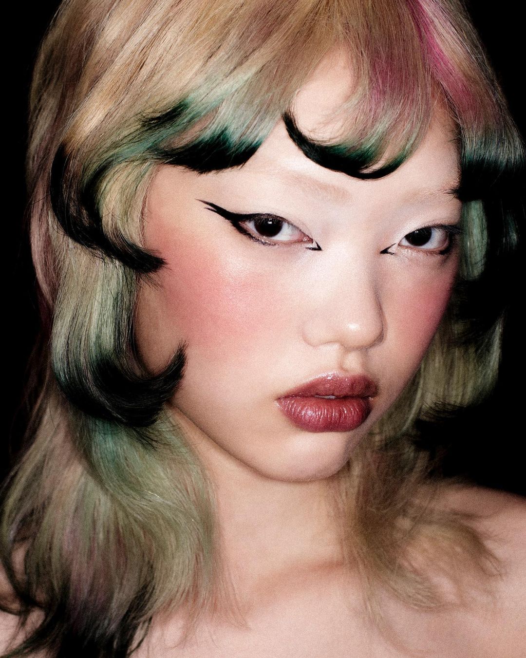 Asian woman's portrait with unique dyed hair and bold winged eyeliner against a dark background, showcasing beauty and fashion.