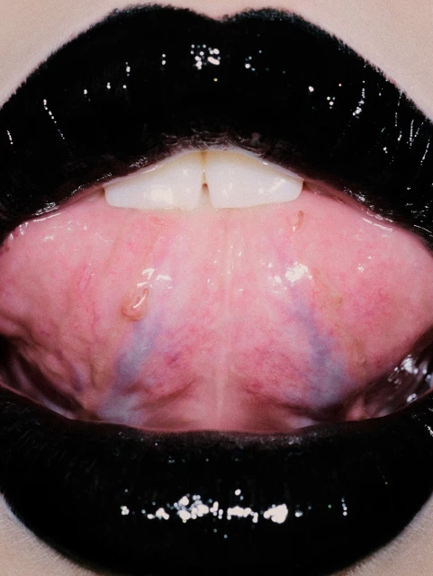 Extreme close-up of a person's mouth featuring glossy black lipstick on their lips, and focusing on the front teeth and pink interior of mouth.