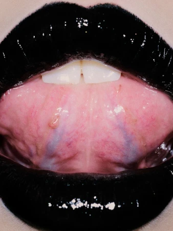 Extreme close-up of a person's mouth featuring glossy black lipstick on their lips, and focusing on the front teeth and pink interior of mouth.