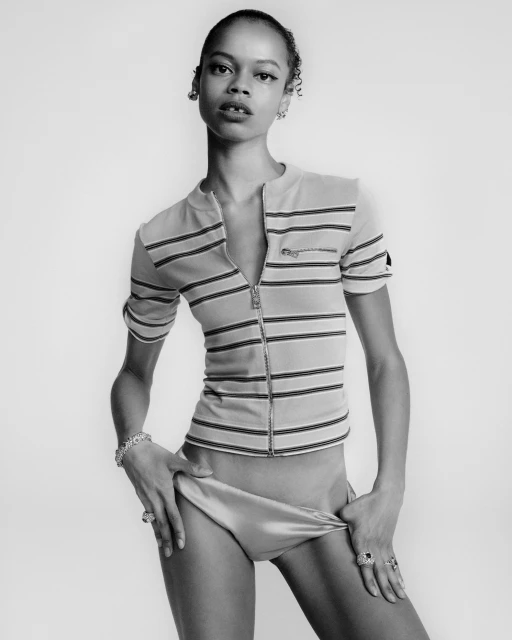 A young woman poses in a striped top and bottoms against a white background, displaying fashion and jewelry in a studio setting.