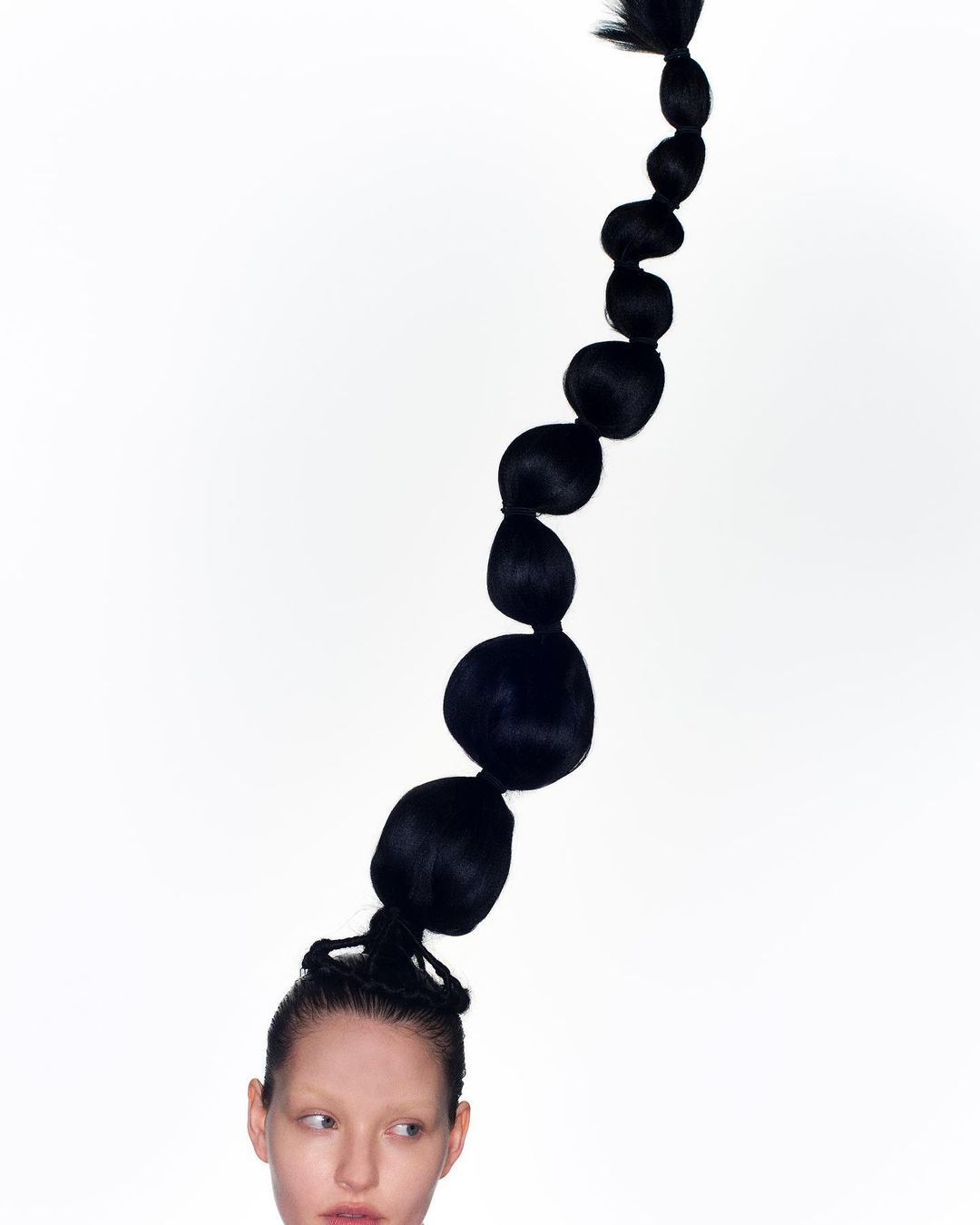 Headshot of a fair-skinned person with black sculptural ponytail hairstyle on a white background, creating a minimalist, surreal portrait.