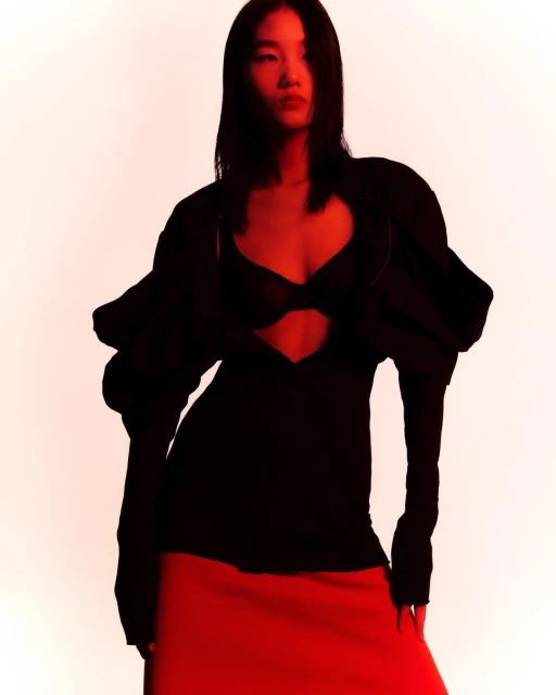 Portrait of an Asian woman in a black top with puffed sleeves and a red skirt against a red-lit white backdrop.
