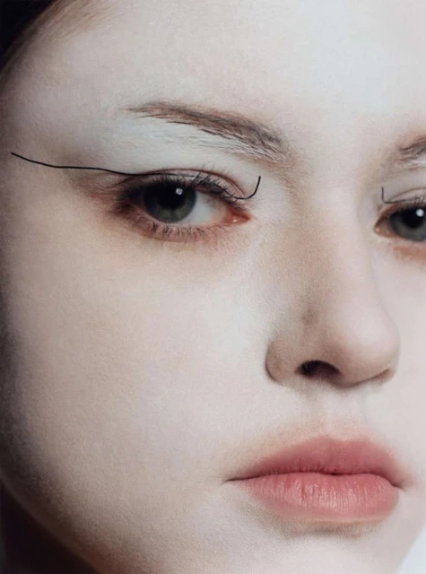 Close-up portrait of a woman's face with white face makeup and black graphic eyeliner, highlighting makeup artistry and facial features.