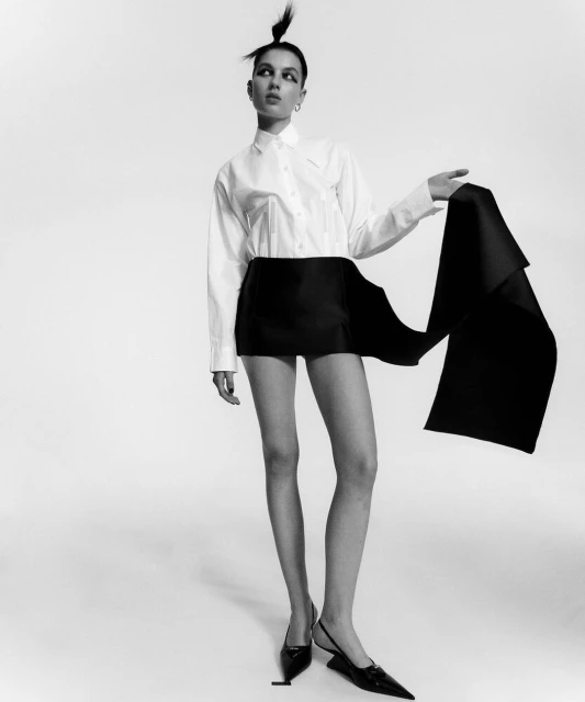 Elegant young adult woman poses in a white button-down shirt, black mini skirt, and slingback heels against a white backdrop.