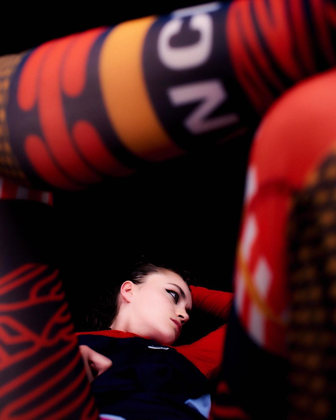Young woman with dark eye makeup lies between orange and black patterned poles in a surreal, high-contrast, studio fashion photo.