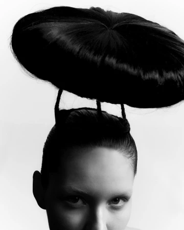 Close-up of a person with an avant-garde black sculpted hair headpiece supported by thin braided strands against a white background.