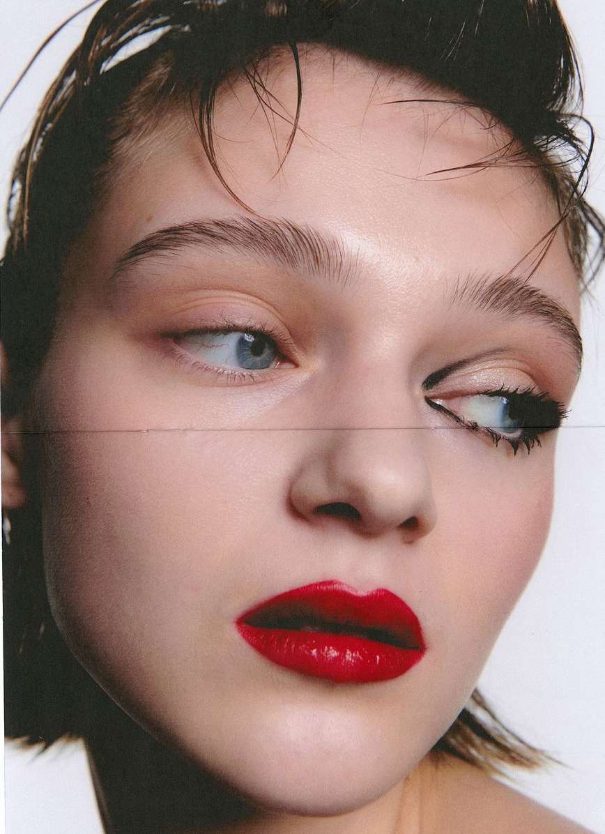 Close-up portrait of a woman with wet hair, bold red lipstick, and graphic black eyeliner against a plain white background.