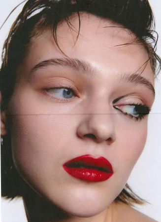 Close-up portrait of a woman with wet hair, bold red lipstick, and graphic black eyeliner against a plain white background.
