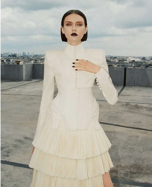 A slender woman with dark makeup posing on a rooftop in a white tiered dress and matching bolero jacket against a cityscape backdrop.