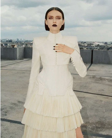 A slender woman with dark makeup posing on a rooftop in a white tiered dress and matching bolero jacket against a cityscape backdrop.