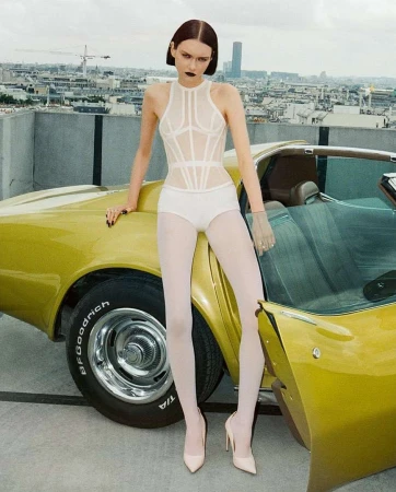 A slender adult female model poses in front of a yellow convertible car on a rooftop with a city skyline backdrop, wearing white lingerie.