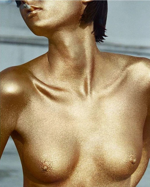 Portrait of a nude adult female covered in gold paint, smoking a cigarette. High-key studio lighting and a neutral grey background.