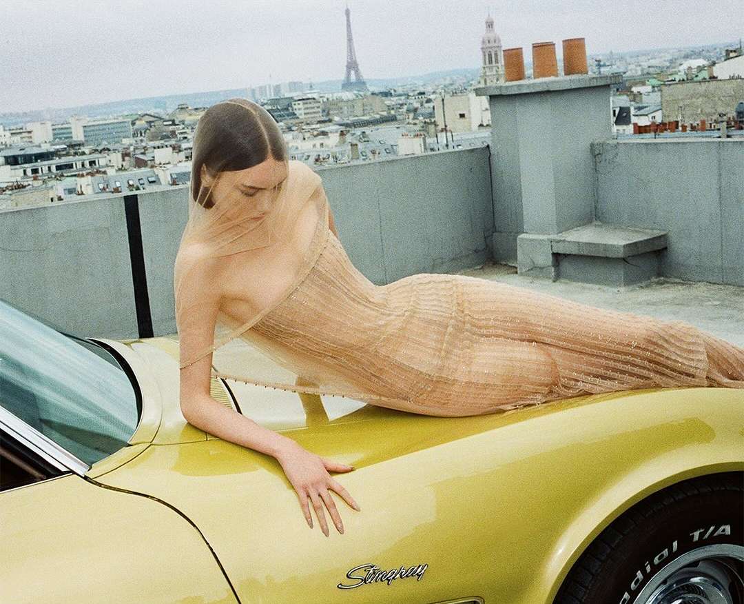 A model in sheer dress lies on a yellow vintage car with the Eiffel Tower in the background in Paris, France, on a rooftop.