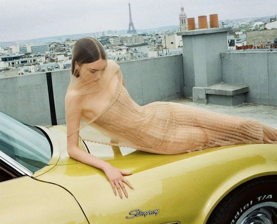A model in sheer dress lies on a yellow vintage car with the Eiffel Tower in the background in Paris, France, on a rooftop.