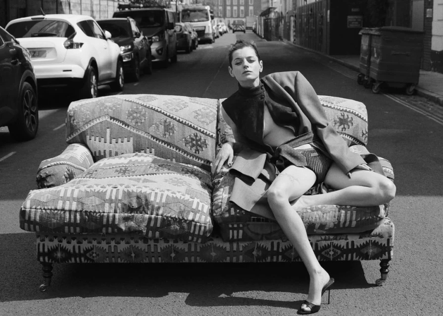 Monochrome fashion shot of a woman in a blazer and heels seated on an ornate couch in a city street, with cars parked nearby.