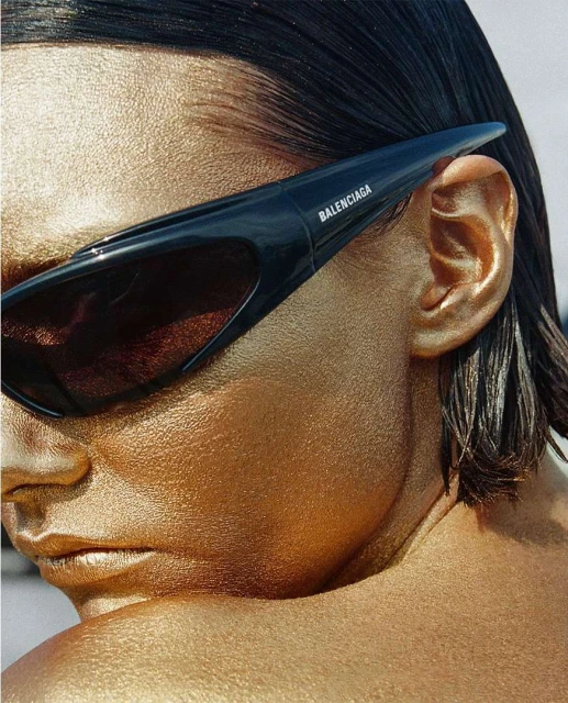Close-up of a woman's face with gold body paint wearing black Balenciaga sunglasses against a neutral background, beauty portrait.