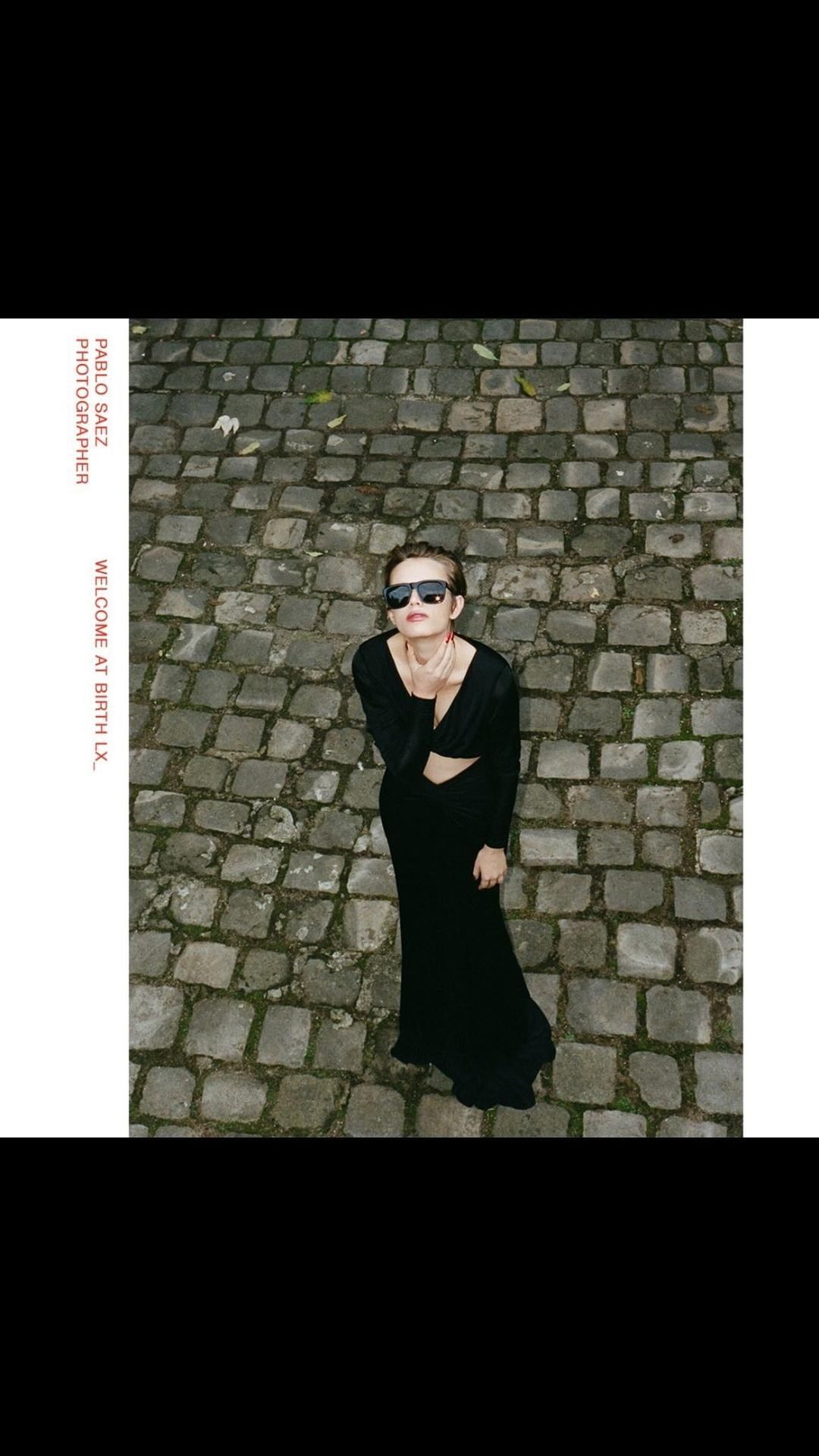 Woman in long black dress and sunglasses poses on cobblestone street. Overhead angle captures fashion against textured stone.