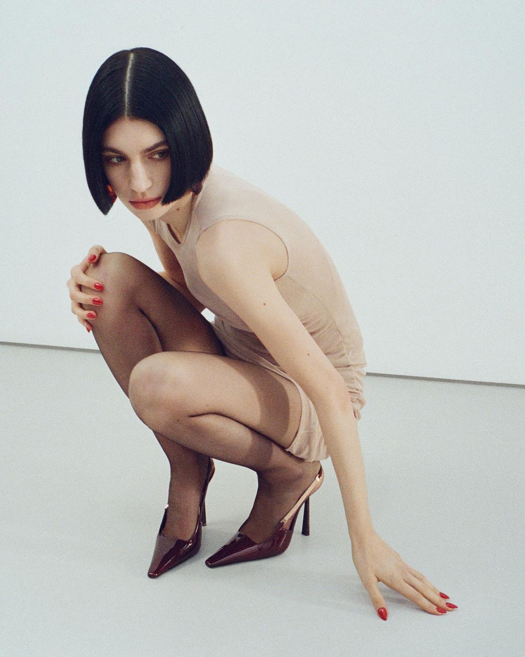 Woman with a bob haircut poses in a beige dress, black tights, and heels against a clean, white backdrop in a studio setting.