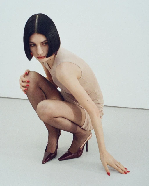 Woman with a bob haircut poses in a beige dress, black tights, and heels against a clean, white backdrop in a studio setting.