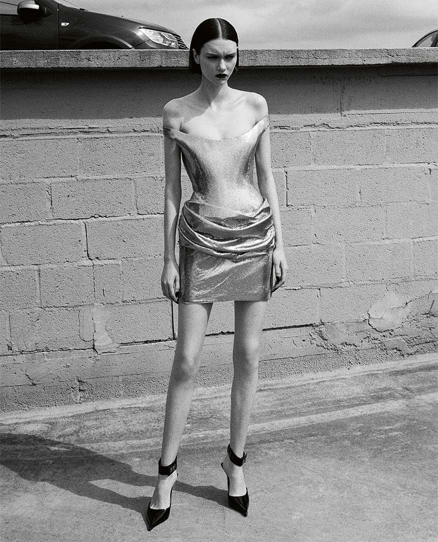 Black and white portrait of a fashion model posing in a metallic mini-dress against a brick wall, highlighting fashion and urban setting.