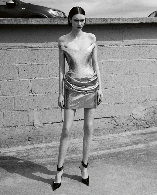 Black and white portrait of a fashion model posing in a metallic mini-dress against a brick wall, highlighting fashion and urban setting.