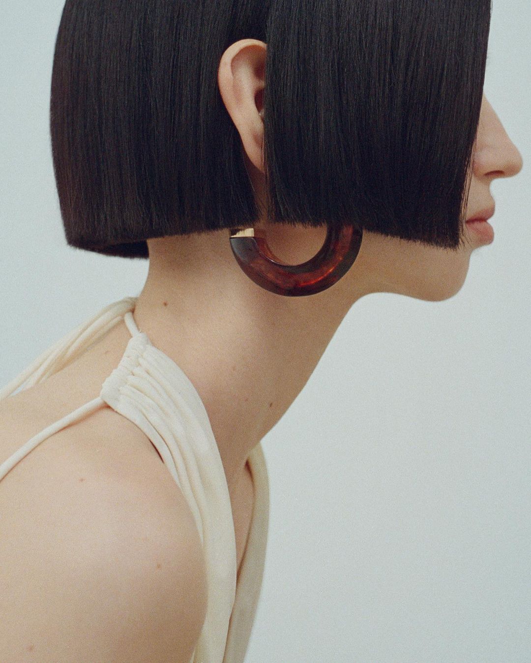 Profile view of a young woman with a dark bob haircut wearing a hoop earring against a clean, neutral background.