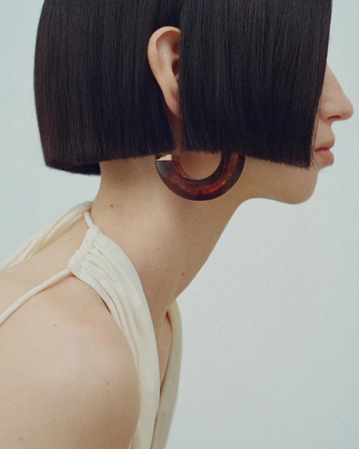Profile view of a young woman with a dark bob haircut wearing a hoop earring against a clean, neutral background.