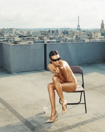 Model with metallic gold skin wearing sunglasses seated on a folding chair on a Paris rooftop overlooking the city skyline