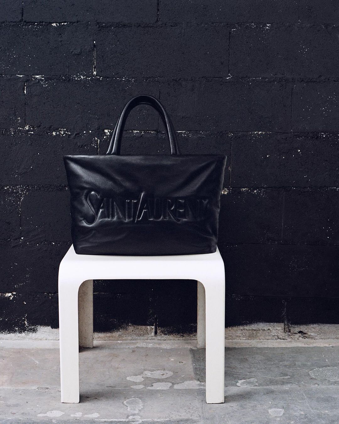 Black Saint Laurent tote bag with embossed logo on a white stool against a dark brick wall backdrop in a minimalist studio shot.