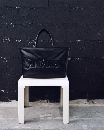 A black leather Saint Laurent tote bag resting on a white plastic stool against a dark textured brick wall.