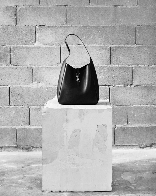 Studio shot of a YSL hobo bag on a textured white podium, set against a geometric concrete block wall, monochrome style.