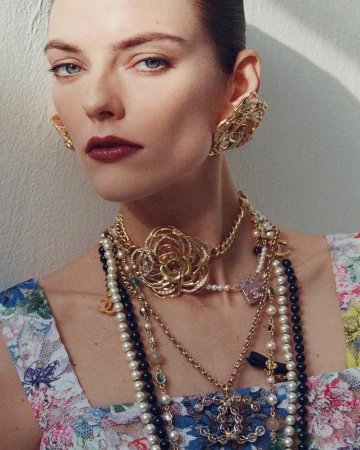 Close-up portrait of a woman wearing layered gold and pearl necklaces with floral earrings against a white wall.