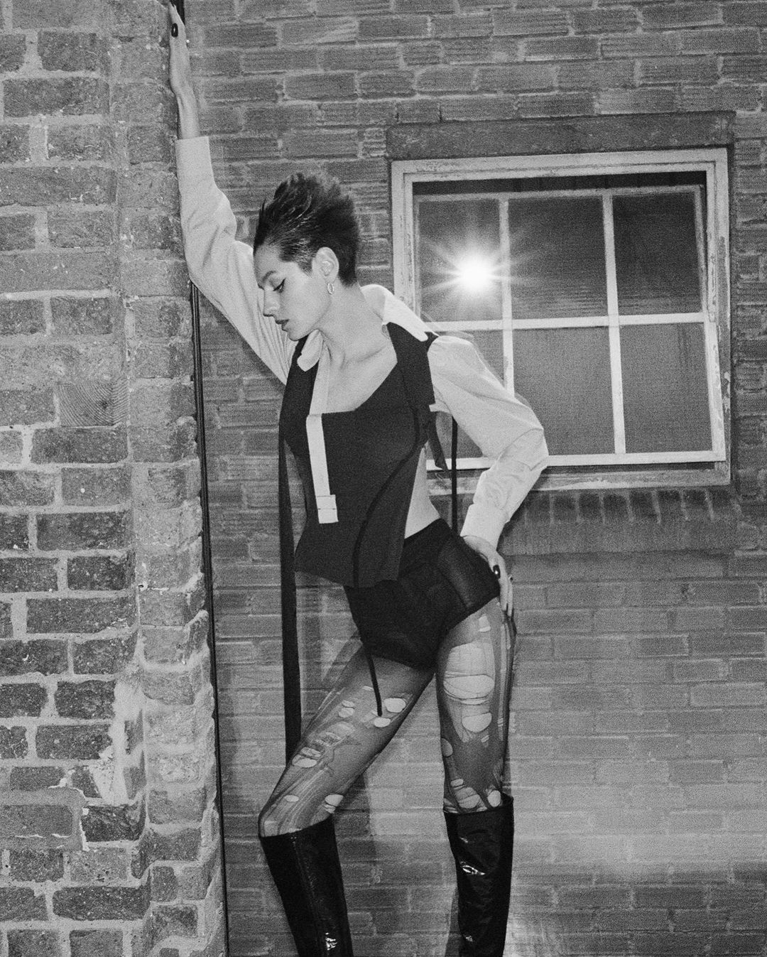 Monochrome fashion shot of a woman in edgy outfit leaning against a brick wall with a window, showcasing a blend of vintage and contemporary styles.