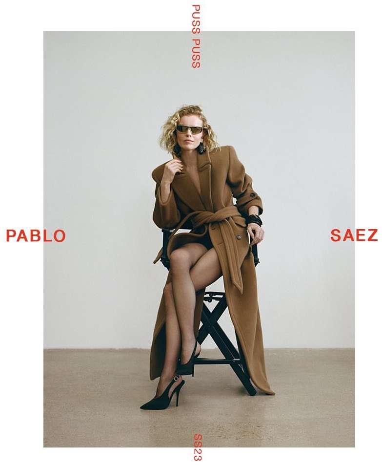 Fashion editorial shot of a woman in a brown coat and black heels sitting on a stool against a white background in a studio setting.