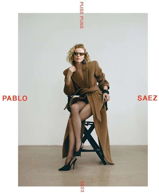 Fashion editorial shot of a woman in a brown coat and black heels sitting on a stool against a white background in a studio setting.