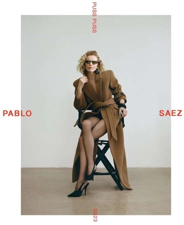Fashion editorial shot of a woman in a brown coat and black heels sitting on a stool against a white background in a studio setting.
