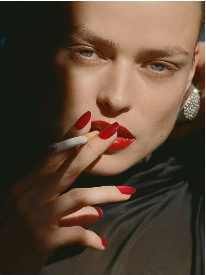 Close-up portrait of a woman with red nails and lipstick smoking a cigarette, wearing sparkly earrings and a black top with dark lighting.