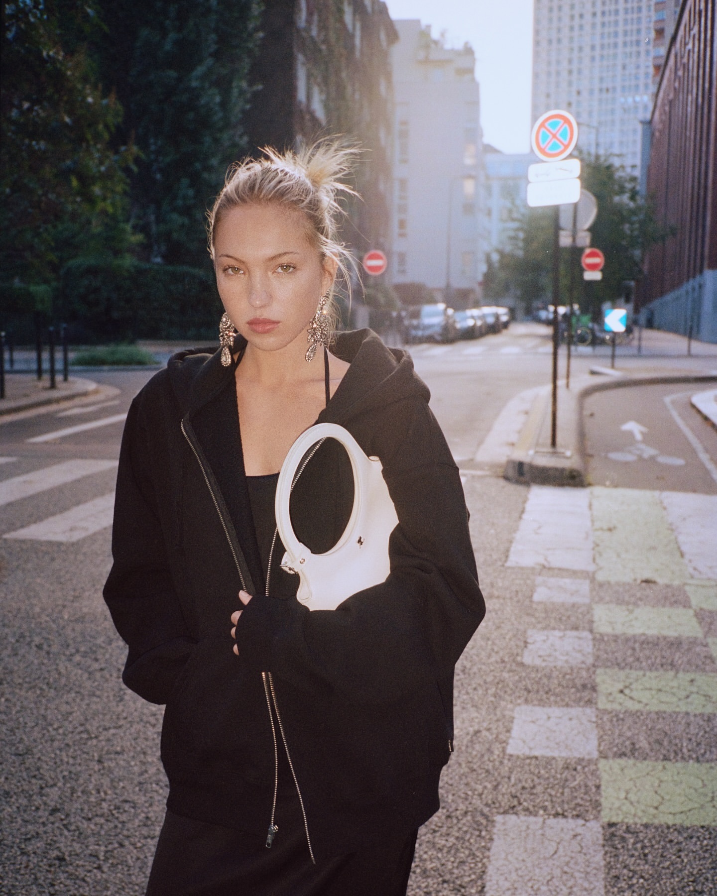 A fashionable young woman in a black zip hoodie, dress, and large earrings poses on a European city street, holding a white handbag.