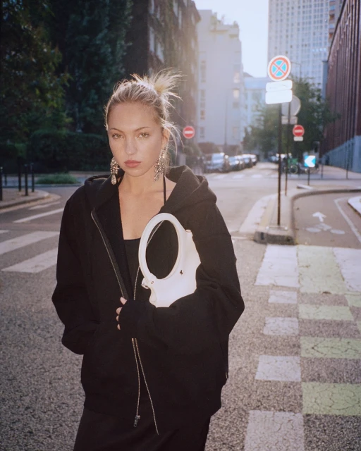 A fashionable young woman in a black zip hoodie, dress, and large earrings poses on a European city street, holding a white handbag.