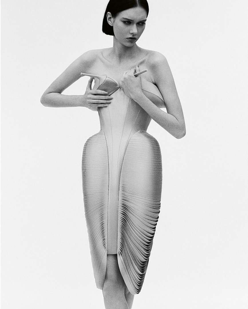 A fashion photo in black and white shows a young woman posing in a sculptural dress, holding high-heeled shoes to her bare chest.