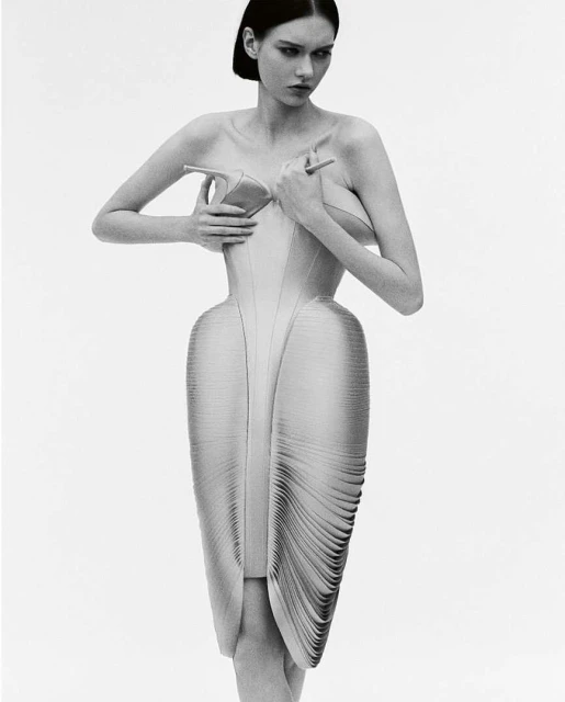 A fashion photo in black and white shows a young woman posing in a sculptural dress, holding high-heeled shoes to her bare chest.