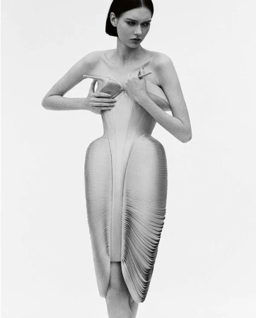 A fashion photo in black and white shows a young woman posing in a sculptural dress, holding high-heeled shoes to her bare chest.