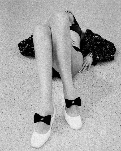 Black and white fashion shot of an adult woman lying on a grey surface, wearing a black bikini and white flats with black bows.