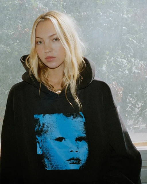 Young adult woman with long blonde hair wearing a black hoodie with a blue child's face graphic, looking at the camera indoors.