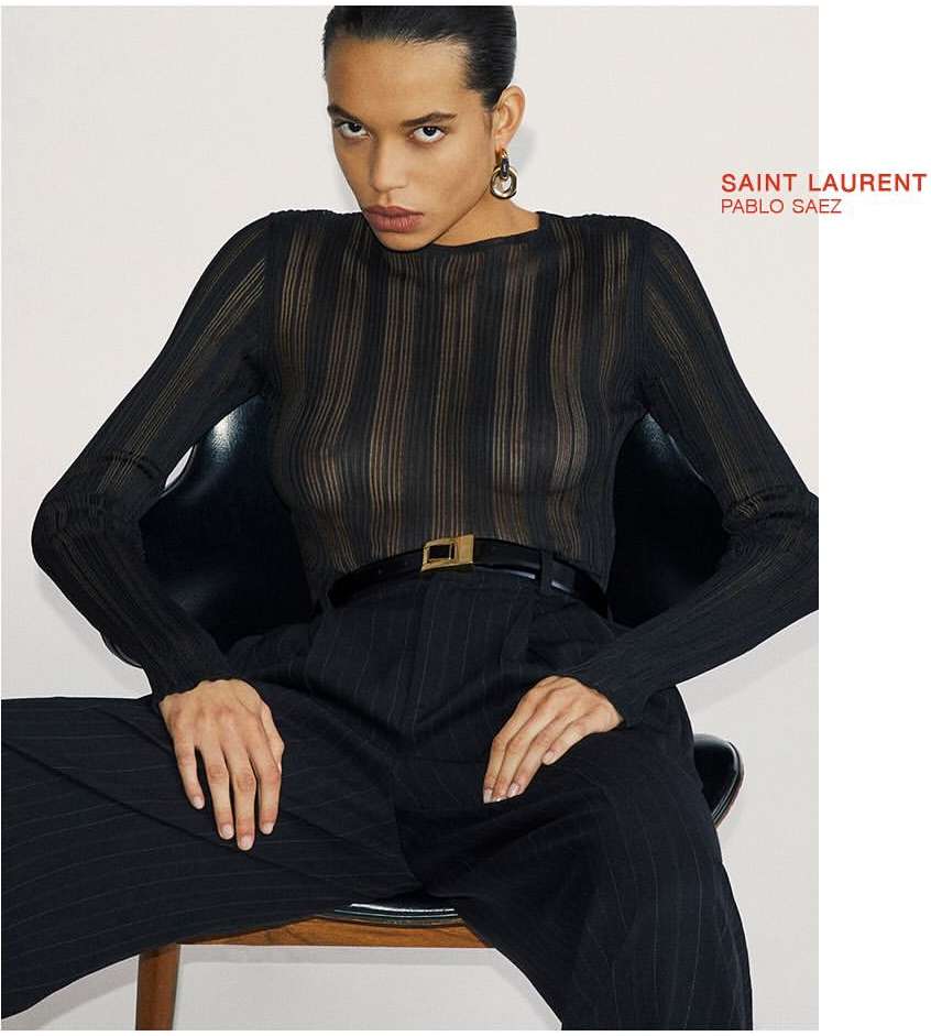 Woman with dark hair and black top and trousers poses against a white wall in a seated position for a fashion editorial.