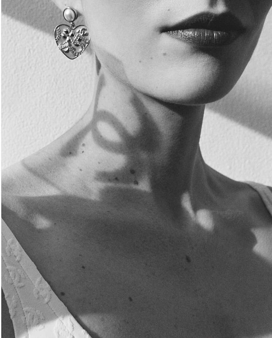 Close-up of a woman's face and neck wearing a heart-shaped earring with white pearl and shadows falling on her skin.