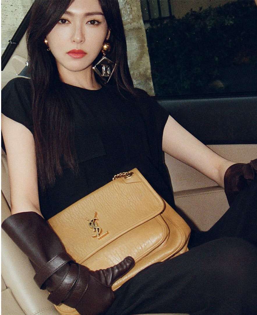 Portrait of an adult woman with long dark hair, wearing large earrings and holding a tan YSL handbag, sitting in a vehicle.