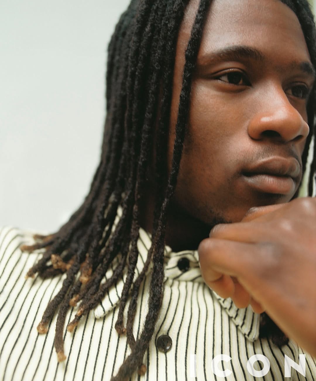 Close-up portrait of an adult Black male with dreadlocks, wearing a striped shirt, his chin resting on his hand, and looking away.